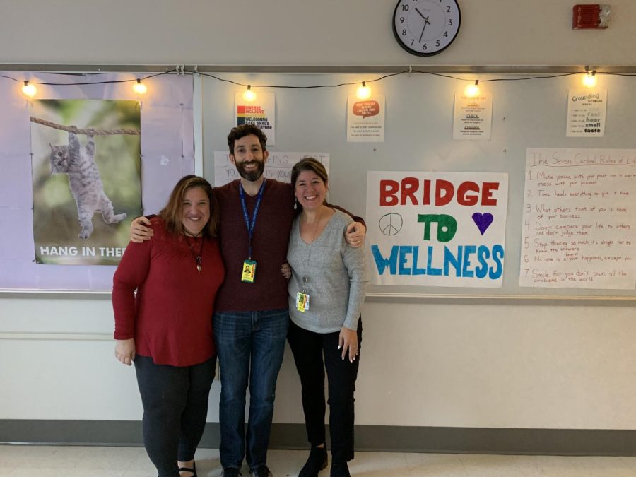 The three members of the Bridge to Wellness team, Maria Bruno, Alex Metral, and Dominic Elliot, stand in front of their Bridge to Wellness room poster.