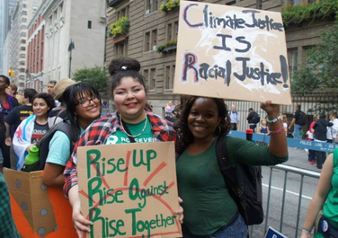 A couple climate activists of color attend and hold signs at a climate change march. One of the signs importantly states Climate Justice IS Racial Justice! 