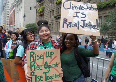 A couple climate activists of color attend and hold signs at a climate change march. One of the signs importantly states "Climate Justice IS Racial Justice!"