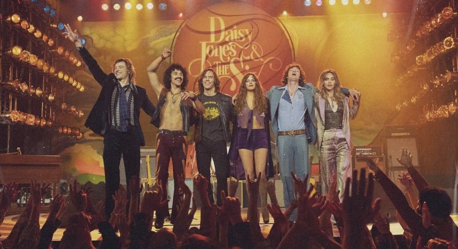 A photo of a stage performance for the fictional band Daisy Jones and the Six. Eddie Roundtree (played by Josh Whitehouse), Warren Rhodes (played by Sebastian Chacon), Billy Dunne (played by Sam Claflin), Daisy Jones (played by Riley Keough), Graham Dunne (played by Will Harrison), and Karen Sirko (played by Suki Waterhouse).
