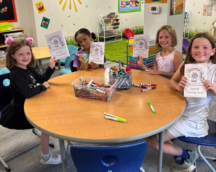 MCPS elementary school students sit around a table with art supplies, posing with their completed coloring books that were created by the WCHS Coloring Club.
